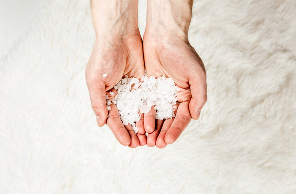 Großaufnahme von Menschenhänden, die Magnesiumchlorid-Vitaminsalzflocken in den Händen halten, isoliert auf weißem, weichem Fellhintergrund. Zutat für ein Fußbad mit Magnesiumsalzflocken. - Foto, Bild