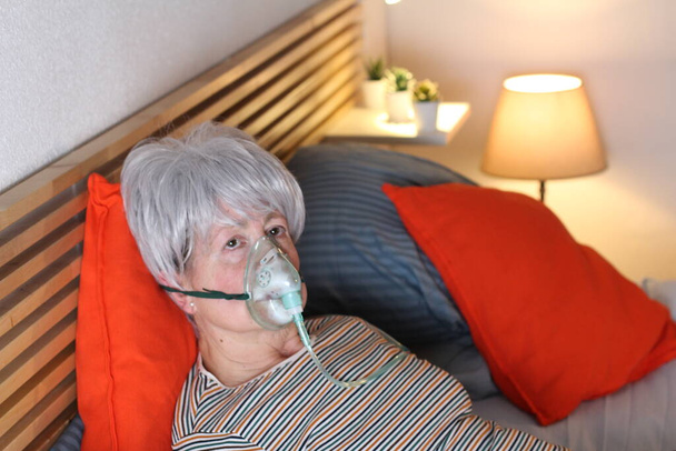 kypsä harmaa tukkainen nainen happinaamarissa rentoutuessaan sängyssä itsensä eristämisen aikana kotona
 - Valokuva, kuva