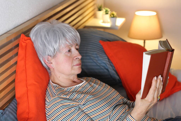 ώριμη γκρίζα μαλλιά γυναίκα ανάγνωση του βιβλίου, ενώ χαλαρώνοντας στο κρεβάτι κατά τη διάρκεια της απομόνωσης στο σπίτι - Φωτογραφία, εικόνα