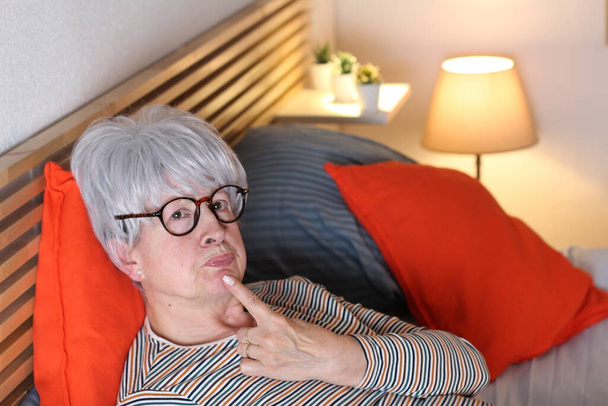 ώριμη γκρίζα μαλλιά γυναίκα αγγίζοντας το πηγούνι της, ενώ χαλαρώνει στο κρεβάτι κατά τη διάρκεια της απομόνωσης στο σπίτι - Φωτογραφία, εικόνα