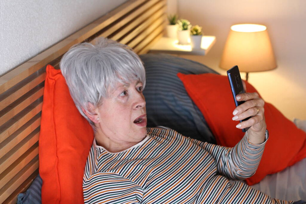 ώριμη γκρίζα μαλλιά γυναίκα χρησιμοποιώντας το smartphone, ενώ χαλαρώνοντας στο κρεβάτι κατά τη διάρκεια της απομόνωσης στο σπίτι - Φωτογραφία, εικόνα