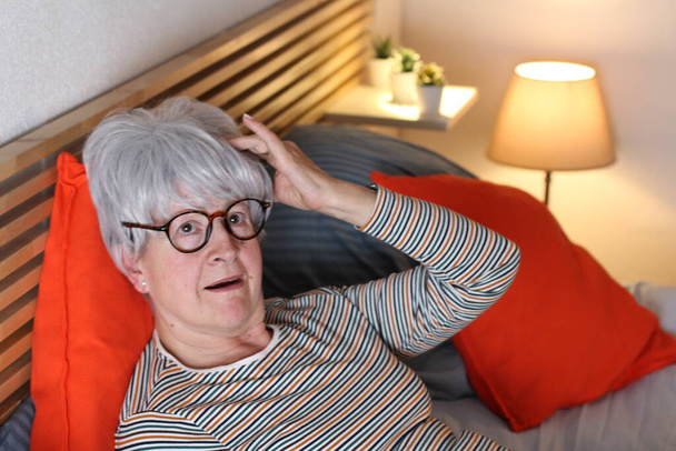 ώριμη γκρίζα μαλλιά γυναίκα που έχει πονοκέφαλο, ενώ χαλαρώνοντας στο κρεβάτι κατά τη διάρκεια της απομόνωσης στο σπίτι - Φωτογραφία, εικόνα