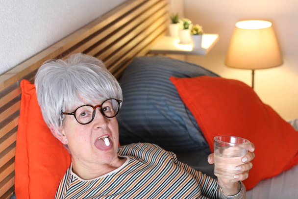 mature femme aux cheveux gris avec un verre d'eau pour prendre ses médicaments tout en se relaxant au lit pendant l'isolement à la maison
 - Photo, image