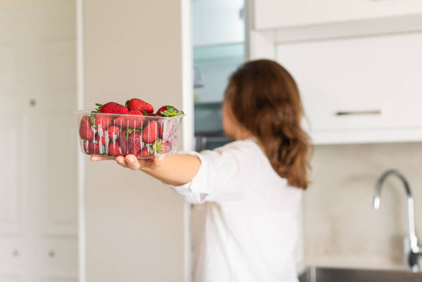 A girl takes out a container of freshly strawberries from the refrigerator. Back view - Foto, afbeelding