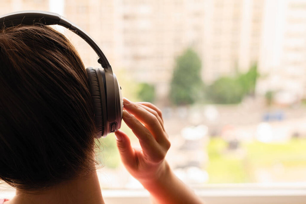 Вид ззаду на самотню молоду жінку, яка слухає музику з навушниками, що стоять біля вікна її квартири. Дівчина дивиться на вулицю
 - Фото, зображення