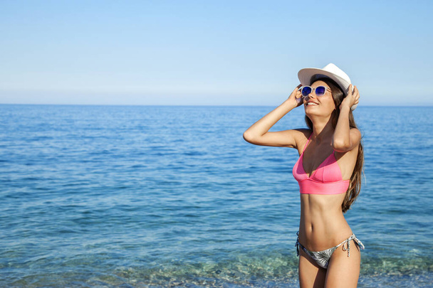 As férias na praia. Mulher bonita quente em chapéu de sol e biquíni de pé com os braços erguidos na cabeça desfrutando de vista para o oceano de praia no dia quente de verão
. - Foto, Imagem