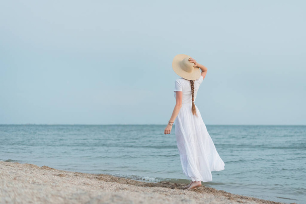 Young woman stands on sandy beach against sea and holding with hand straw hat. - Photo, Image