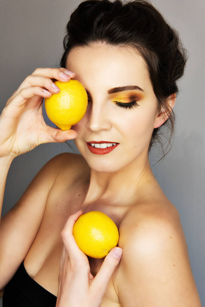 Beautiful girl with strong colorfull makeup, yellow, brown and orange eyeshadows. Woman is holding lemon in front of her one eye. - Photo, Image