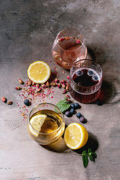 Verschiedene Cocktailgläser mit prickelnden bunten Getränken rosa Rosenchampagner, Apfelwein oder Limonade mit trockenen Rosenblüten, Zitrone, Blaubeeren und Minze. Zutaten oben. Graue Textur Hintergrund. - Foto, Bild
