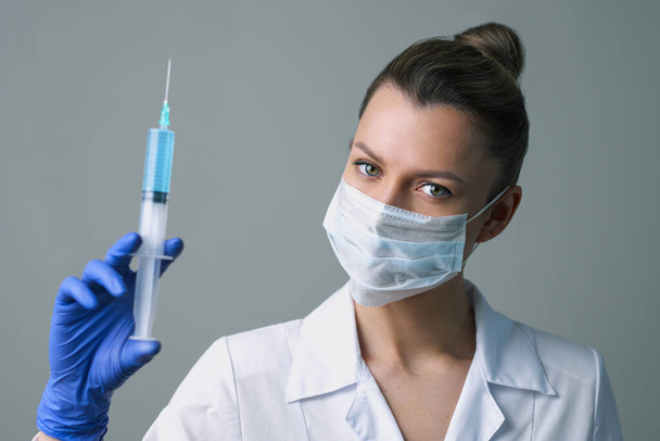 A female medical worker in a white robe and mask holds a syringe. - Valokuva, kuva