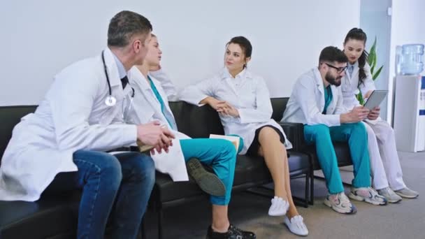 In a modern hospital group of the doctors and nurses have a break time in the stuff room they relaxing a bit discussing and take a tablet to watch something - Materiał filmowy, wideo