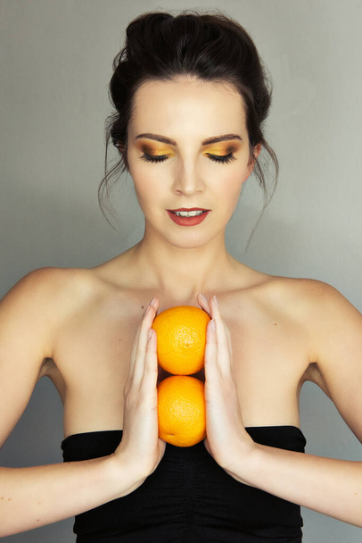 Beautiful girl with strong colorfull makeup, yellow, brown and orange eyeshadows, holding two orange in front of her chest - Φωτογραφία, εικόνα