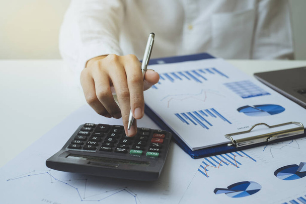 Close-up hand of businessman holding a pen and using a calculator to calculate the numbers, tax and analyze graph chart and business data on the white desk in the office. finance accounting concept. - 写真・画像