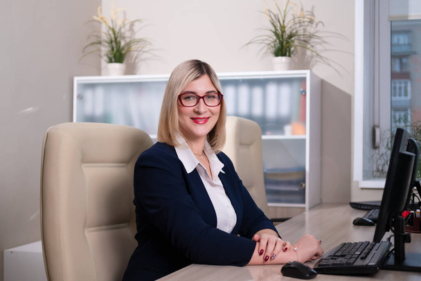 A young blonde woman with glasses is an office worker. Woman sits at the table and works at the computer. - Fotó, kép