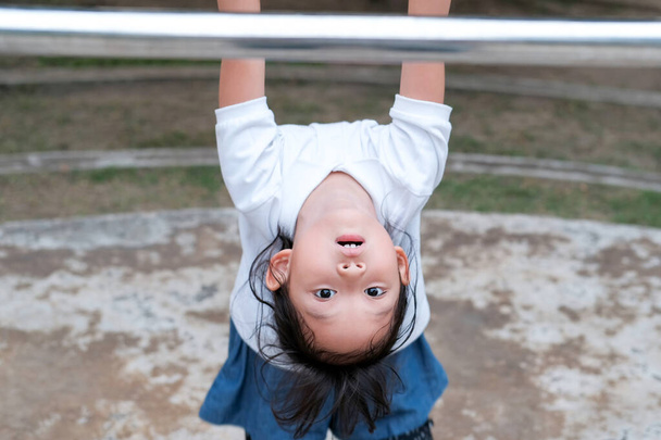 Cute Asian kid hanging on pull-up bar at playground in the village park. Sport outdoor activity for kid. - Photo, Image