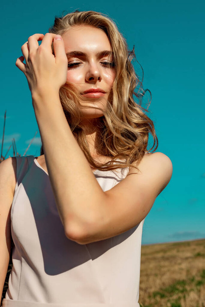 Portrait of a blonde young girl model who stands and poses on a sunny day against a background of haystacks and blue sky enjoying the rays of the sun. - Foto, Bild
