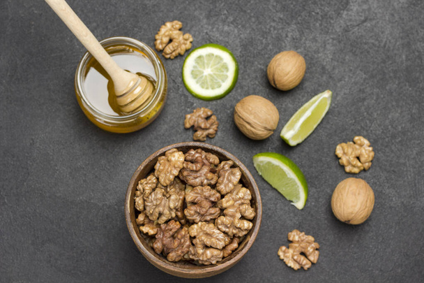 Walnut kernels in wooden bowl, honey in glass jar, whole nuts, lime slices on table. Black background. Flat lay - Fotó, kép