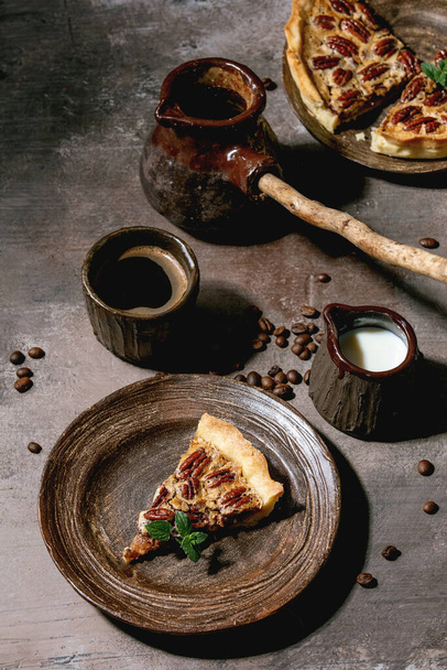 Piece of home baking classic american pecan caramel pie on ceramic plate, coffee cup, cezve, jug of cream over grey texture background. - Fotoğraf, Görsel