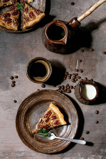 Piece of home baking classic american pecan caramel pie on ceramic plate, coffee cup, cezve, jug of cream over grey texture background. Flat lay, space - Foto, immagini