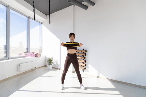 Dziewczyna w dresie trenuje z elastyczną opaską fitness w lekkim studiu rozciągającym. Trening rano w słoneczny dzień - Zdjęcie, obraz