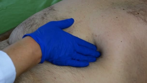 Lekarz dotyka ręką w niebieskiej gumowej rękawiczce żołądka chorego mężczyzny, który leży w łóżku. Zbliżenie. - Materiał filmowy, wideo