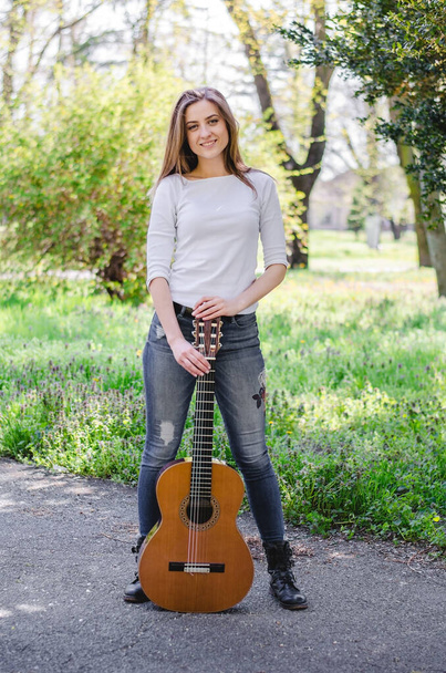 Chica yound de pie y sosteniendo la guitarra en posición vertical en el parque. Sonriendo y mirando a la cámara
. - Foto, Imagen