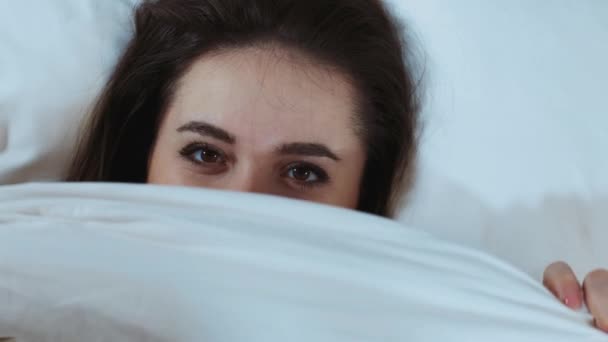 Top view of a cheerful young woman hiding under a soft white blanket, model plus size having fun in bed, looking at the camera, enjoying good morning, resting in bed. View from above. Copy space - Filmati, video