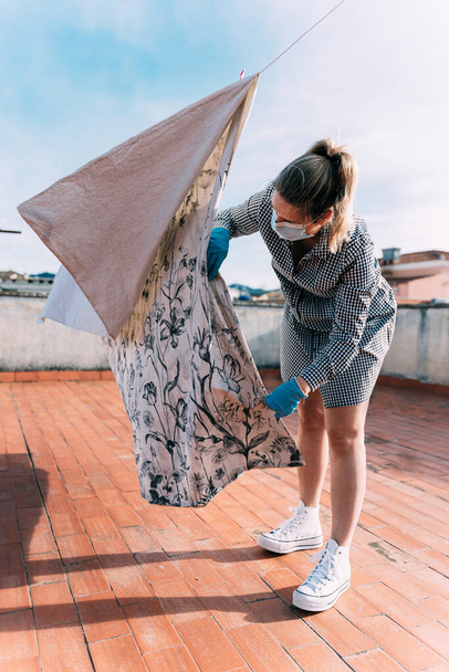 Coronavirus. Woman in quarantine with face mask at home on the roof or balcony, hanging out the washing isolated. Disinfecting bed clothes. Virus prevention and protocols. Stay at home. Confinement - Photo, Image