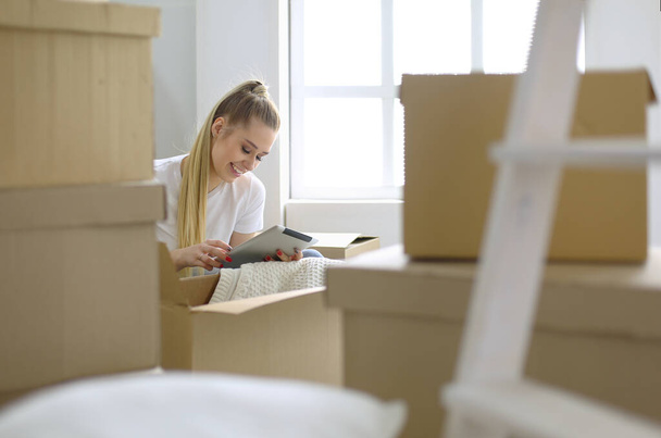 A beautiful single young woman unpacking boxes and moving into a new home. A women look at here tablet - Фото, изображение
