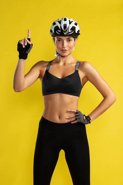 常に自転車のヘルメットを着用してください。黄色の背景に立っている間、上を指してカメラを見て自転車のヘルメットで若いスポーティ女性。スタジオショット - 写真・画像