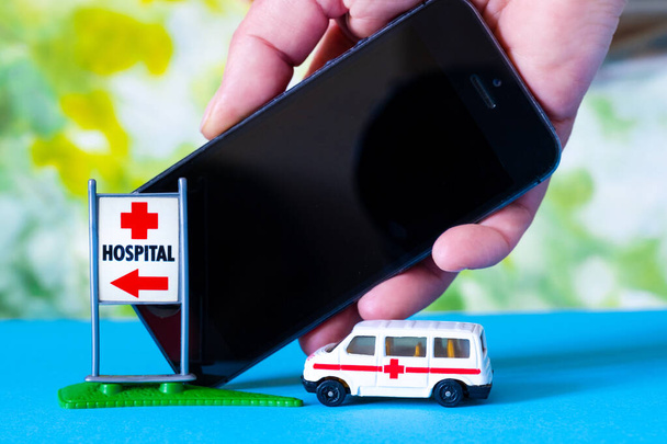 Παιχνίδι ασθενοφόρο αυτοκίνητο και το χέρι με το τηλέφωνο κοντά σημάδι Νοσοκομείο στο μπλε και πράσινο φωτεινό φόντο. - Φωτογραφία, εικόνα