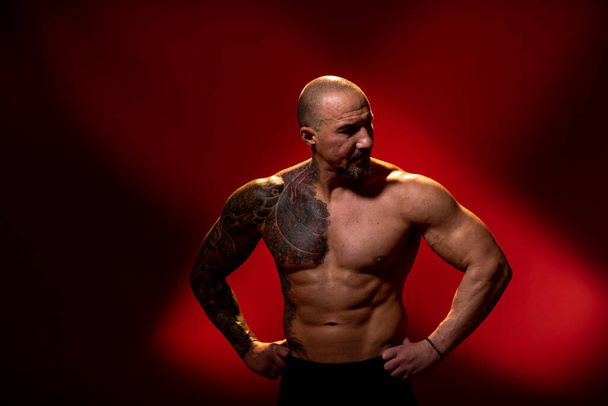 Un sportif avec un tatouage posé sur fond rouge. Entraîneur de fitness avec torse nu, debout dans le studio. Bodybuilder détourner les yeux
. - Photo, image