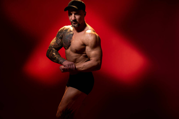 Homem musculoso e forte em pose de musculação típica. Fisiculturista com tatuagem na tampa no fundo vermelho. Vista lateral, espaço de cópia
. - Foto, Imagem