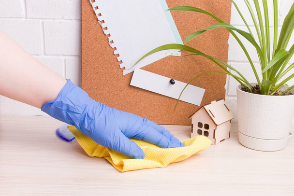 a female hand in a blue rubber glove wipes a desktop with a yellow rag, a flower in a pot and a cork board with notes on the background, a model of a wooden house, work from hom - Fotografie, Obrázek