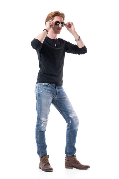 Homem bonito jovem fresco com cabelo vermelho vestindo jeans rasgado e camisa preta experimentando em óculos de sol. Corpo inteiro isolado em fundo branco
.  - Foto, Imagem