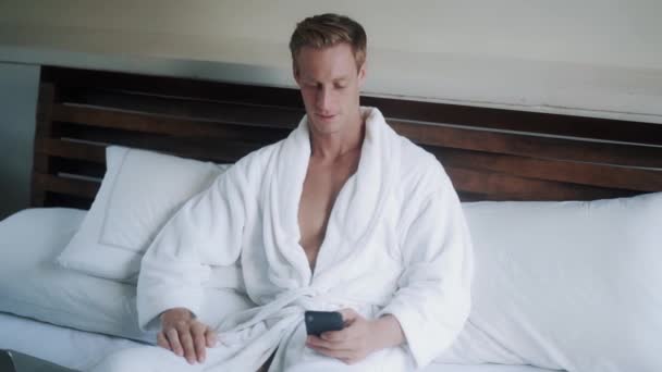 Handsome blond guy in white bathrobe use mobile phone and answers calling in bed - Filmmaterial, Video