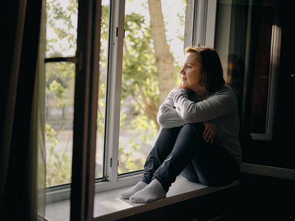 Una bella giovane donna è triste seduta su una finestra della sua casa. Quarantena dovuta alla pandemia di coronavirus
. - Foto, immagini