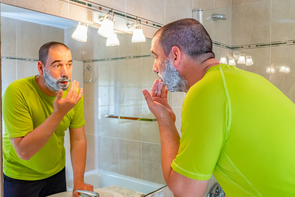 Мужчина с бритой головой и в фисташково-зеленой рубашке бреет бороду перед зеркалом
 - Фото, изображение