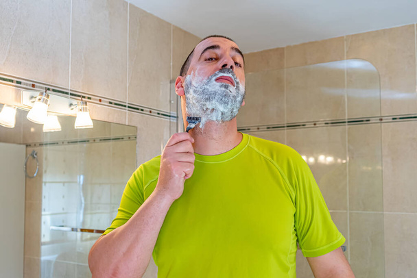 Man with a shaved head and wearing a pistachio green shirt shaving his beard in front of the mirror - Photo, Image