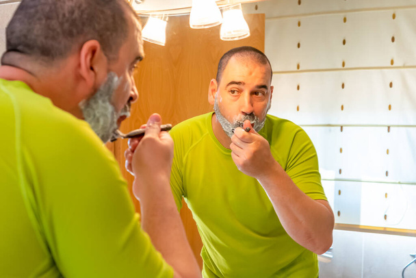 Borotvált fejű, pisztácia zöld inges férfi, aki a tükör előtt borotválja a szakállát. - Fotó, kép
