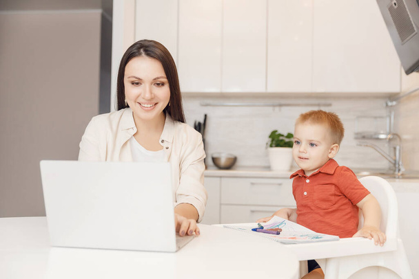 Nő szülési szabadságon dolgozik otthon online laptoppal a konyhában kisgyerekkel. Fogantatás anya munka közben karantén elkülönítés során Covid-19 - Fotó, kép