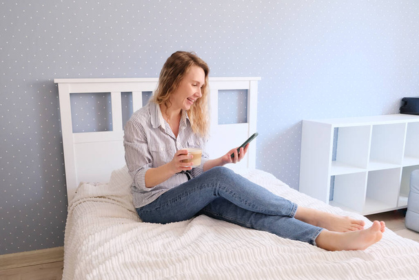 Mladá žena pije kávu a mluví na smartphone, zatímco sedí na posteli - Fotografie, Obrázek