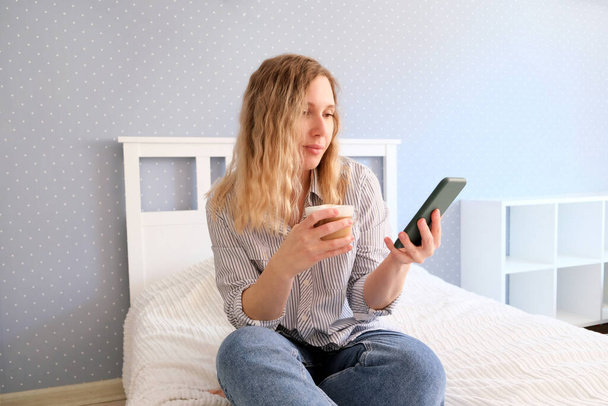 若い女性はコーヒーを飲み、ベッドに座っている間にスマートフォンで話します - 写真・画像