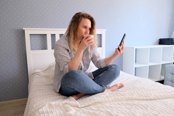 Jeune femme boit du café et parle sur le smartphone tout en étant assis sur le lit
 - Photo, image