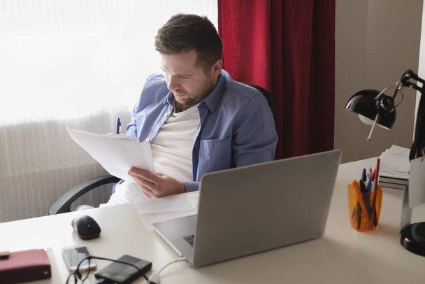 Кавказький чоловік працює вдома на ноутбуці, читаючи документи.. - Фото, зображення