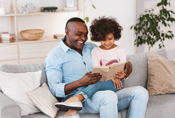 Окремі заходи. Цікава афроамериканська дівчинка, яка читає книжку разом з дідусем у вітальні. - Фото, зображення