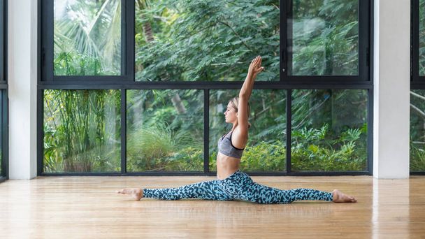 Panoramic and side view of young adult sporty girl doing yoga, raised hands up, sitting in split, making stretching and flexibility training against nature and plants on background - Photo, Image