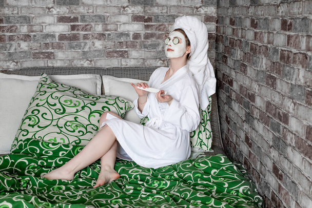 menina bonito em um roupão de banho de waffle branco e toalha na cabeça fazendo manicure com pepinos em seus olhos
 - Foto, Imagem