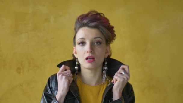 Glamouröse Dame in Punk-Rock-Party-Kleidung mit dunkelrosa Haaren in lässigem Kleid, schwarzer Lederjacke und langen Ohrringen auf gelbem Hintergrund - Filmmaterial, Video