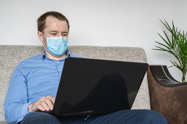 Een zakenman die thuis werkt draagt een beschermend masker. Blanke volwassen man in een blauw shirt zittend op een bank, werkend met een laptop. Werk vanuit huis tijdens quarantaine. Coronavirus. - Foto, afbeelding